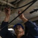 A U.S. Navy sailor identified as Roberto Martinez, CS-3, is performing maintenance work overhead, using tools on cables or conduits attached to a ship's ceiling. He is wearing standard Navy working uniform (NWU), a blue hard hat, and safety glasses. The setting appears to be an interior compartment of a naval vessel.