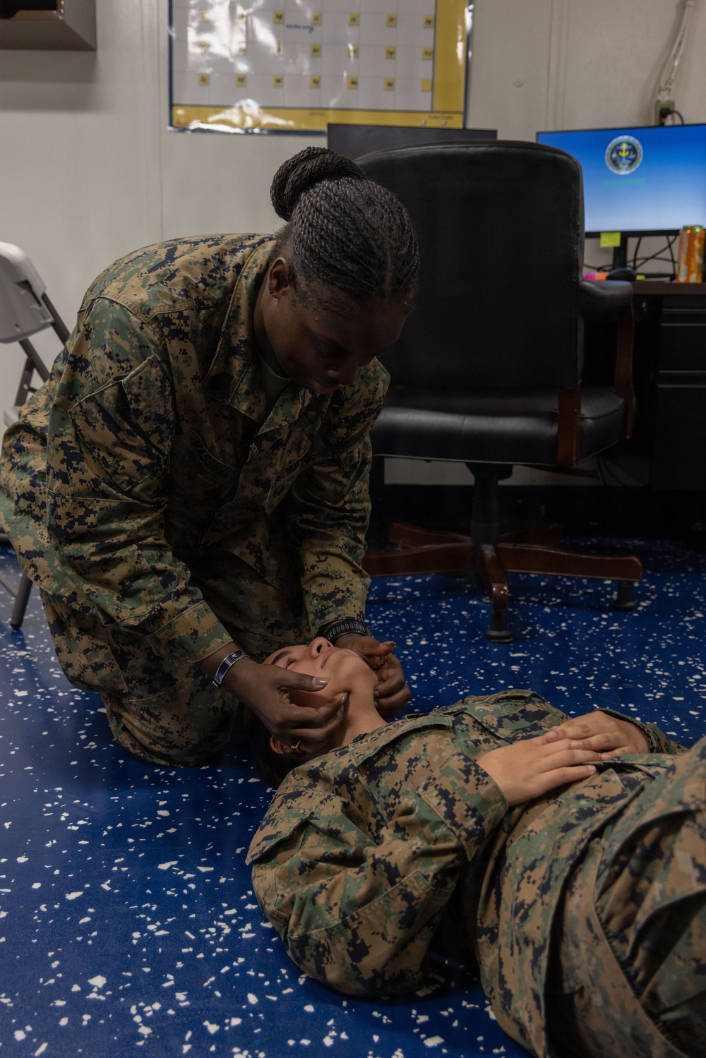 Two military personnel in MARPAT (Marine pattern) camouflage uniforms are conducting what appears to be a first aid or CPR training exercise. One service member is kneeling and performing a jaw-thrust or head-tilt/chin-lift maneuver on a second service member lying on the floor. The setting appears to be an indoor military office or training space.