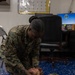 Two military personnel in MARPAT (Marine pattern) camouflage uniforms are conducting what appears to be a first aid or CPR training exercise. One service member is kneeling and performing a jaw-thrust or head-tilt/chin-lift maneuver on a second service member lying on the floor. The setting appears to be an indoor military office or training space.