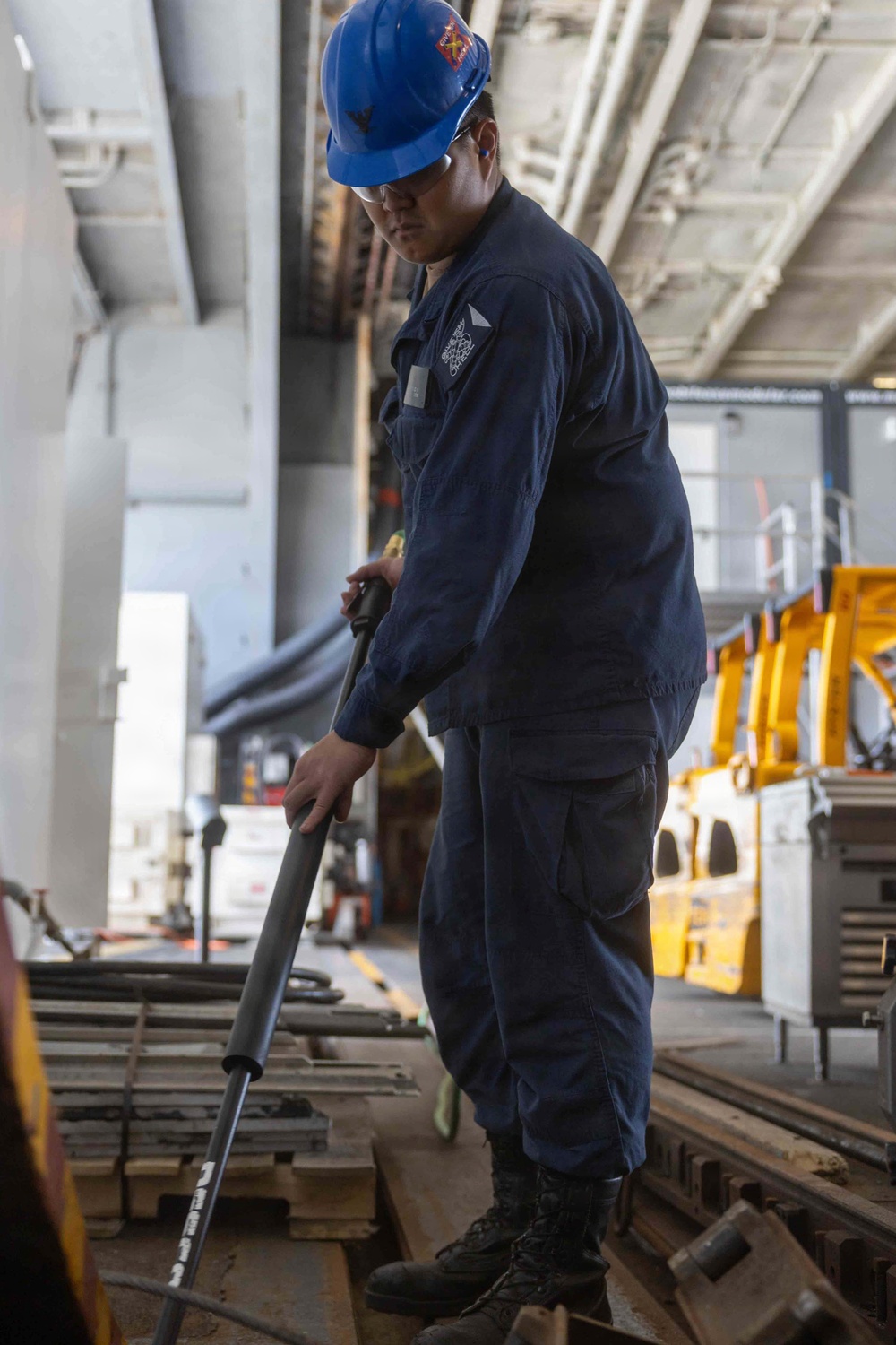 A worker in navy blue coveralls and a blue hard hat is standing in what appears to be a large industrial or military hangar bay, handling a long metal rod or tool. The person is wearing safety equipment including ear protection and safety glasses, and is looking downward at equipment or materials on the floor. Yellow heavy machinery is visible in the background.