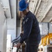 A worker in navy blue coveralls and a blue hard hat is standing in what appears to be a large industrial or military hangar bay, handling a long metal rod or tool. The person is wearing safety equipment including ear protection and safety glasses, and is looking downward at equipment or materials on the floor. Yellow heavy machinery is visible in the background.
