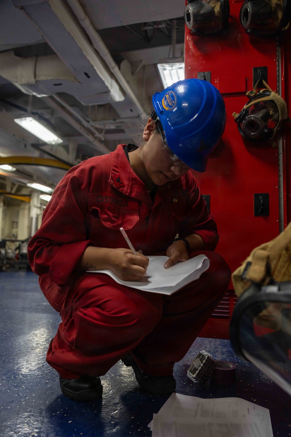 A person wearing red coveralls and a blue hard hat is crouched down writing on paper while conducting what appears to be an inspection or documentation task aboard a naval vessel. The individual's name tag reads 'CASTANEDA' and they are positioned next to red firefighting or damage control equipment. The industrial interior setting with blue-painted deck, overhead piping, and mounted breathing apparatus is consistent with a U.S. Navy ship's interior compartment.