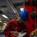 A person wearing red coveralls and a blue hard hat is crouched down writing on paper while conducting what appears to be an inspection or documentation task aboard a naval vessel. The individual's name tag reads 'CASTANEDA' and they are positioned next to red firefighting or damage control equipment. The industrial interior setting with blue-painted deck, overhead piping, and mounted breathing apparatus is consistent with a U.S. Navy ship's interior compartment.