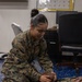 A U.S. Marine in MARPAT camouflage uniform is kneeling on a blue speckled mat, performing what appears to be a first aid or CPR training exercise on another Marine who is lying on their back. The kneeling Marine, whose name tape reads 'CHAPARRO' or similar, is positioning the head of the supine Marine whose name tape reads 'MASSINGILL'. The setting appears to be an indoor training room or office space.