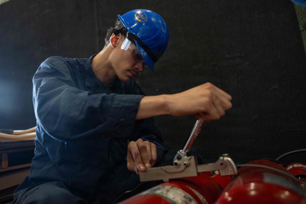 A young male worker wearing a blue hard hat and navy coveralls is using a wrench to service or inspect red fire extinguishers. He appears focused on tightening or loosening a valve or fitting on the equipment. The setting appears to be an industrial or maritime workspace with a dark background.
