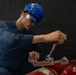 A young male worker wearing a blue hard hat and navy coveralls is using a wrench to service or inspect red fire extinguishers. He appears focused on tightening or loosening a valve or fitting on the equipment. The setting appears to be an industrial or maritime workspace with a dark background.
