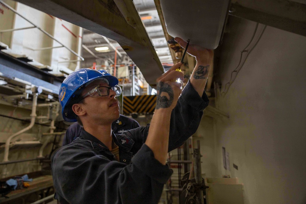 A male worker wearing a blue hard hat and safety glasses is performing maintenance or inspection work on overhead equipment in an industrial facility. He is using a screwdriver and a small flashlight to work on what appears to be a panel or fixture mounted above him. The environment appears to be a large industrial space, possibly a shipyard, factory, or hangar.