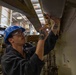 A male worker wearing a blue hard hat and safety glasses is performing maintenance or inspection work on overhead equipment in an industrial facility. He is using a screwdriver and a small flashlight to work on what appears to be a panel or fixture mounted above him. The environment appears to be a large industrial space, possibly a shipyard, factory, or hangar.