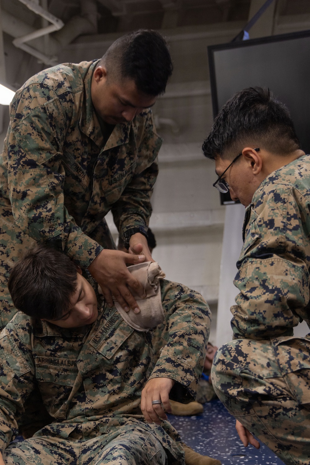 Three U.S. Marines in MARPAT digital camouflage uniforms are engaged in what appears to be a combat casualty care or first aid training exercise. Two Marines are applying what appears to be a bandage or pressure dressing to the head/neck area of a third Marine who is seated on the floor acting as a simulated casualty. The setting appears to be an indoor military facility, possibly aboard a naval vessel.