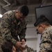 Three U.S. Marines in MARPAT digital camouflage uniforms are engaged in what appears to be a combat casualty care or first aid training exercise. Two Marines are applying what appears to be a bandage or pressure dressing to the head/neck area of a third Marine who is seated on the floor acting as a simulated casualty. The setting appears to be an indoor military facility, possibly aboard a naval vessel.