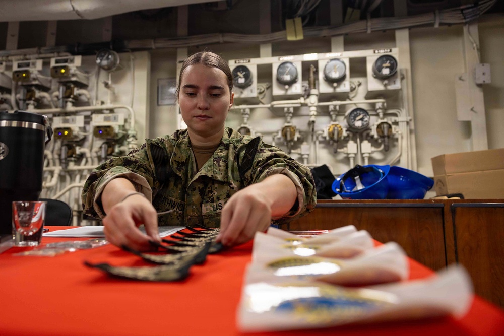 A U.S. Navy sailor in NWU Type III (woodland camouflage) uniform is seated at a red-covered table aboard what appears to be a naval vessel, handling military insignia or patches. The background features a panel of pressure gauges, valves, and piping consistent with a shipboard engineering or mechanical space. Various items including patches, medals, and documents are spread across the table.