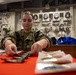 A U.S. Navy sailor in NWU Type III (woodland camouflage) uniform is seated at a red-covered table aboard what appears to be a naval vessel, handling military insignia or patches. The background features a panel of pressure gauges, valves, and piping consistent with a shipboard engineering or mechanical space. Various items including patches, medals, and documents are spread across the table.