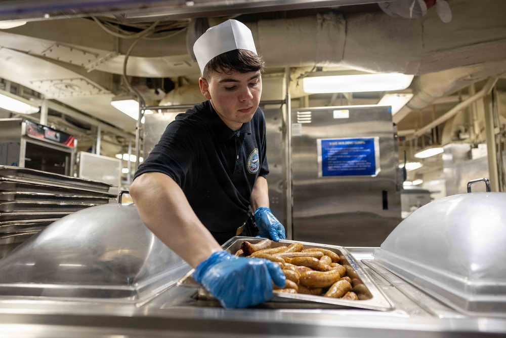 A young food service worker wearing a white paper chef's hat and dark navy uniform with an emblem is placing a tray of cooked sausages into a steam table serving station. The setting appears to be a military galley or institutional kitchen, identifiable by the stainless steel equipment, overhead piping, and a visible 'Safe Holding Temps' notice posted on the wall. The worker is wearing blue disposable gloves and handling the food with care.