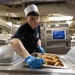 A young food service worker wearing a white paper chef's hat and dark navy uniform with an emblem is placing a tray of cooked sausages into a steam table serving station. The setting appears to be a military galley or institutional kitchen, identifiable by the stainless steel equipment, overhead piping, and a visible 'Safe Holding Temps' notice posted on the wall. The worker is wearing blue disposable gloves and handling the food with care.