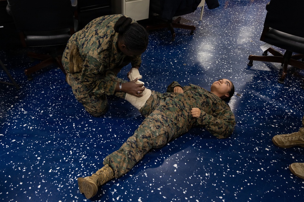 Two military personnel in MARPAT camouflage uniforms are engaged in a first aid or combat casualty care training exercise on a blue speckled floor. One service member is kneeling and applying a bandage or wound dressing to the midsection/abdomen area of another service member who is lying on the floor. The setting appears to be an indoor facility, likely a classroom or briefing room, with chairs visible in the background.