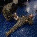 Two military personnel in MARPAT camouflage uniforms are engaged in a first aid or combat casualty care training exercise on a blue speckled floor. One service member is kneeling and applying a bandage or wound dressing to the midsection/abdomen area of another service member who is lying on the floor. The setting appears to be an indoor facility, likely a classroom or briefing room, with chairs visible in the background.