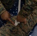 A person wearing USMC MARPAT (Marine Pattern) digital camouflage uniform is applying what appears to be gauze or a wound packing material to a simulated injury on a leg or limb also dressed in MARPAT uniform. The setting appears to be an indoor training environment with a blue floor mat scattered with white foam debris. This appears to be a tactical combat casualty care (TCCC) or first aid training exercise.