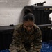 A female U.S. Marine in MARPAT woodland digital camouflage uniform is kneeling on a blue mat, cleaning or treating a second camouflage uniform laid flat in front of her using a white cloth or rag. The setting appears to be an indoor storage or utility room with black plastic storage containers visible in the background. The image captures a routine uniform maintenance task.