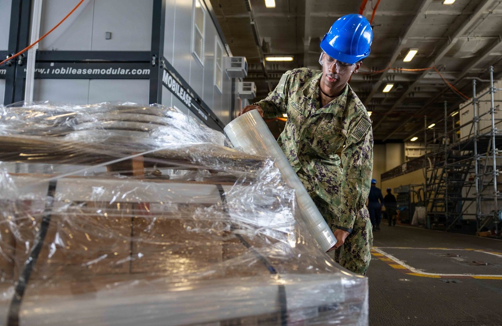 A U.S. Navy sailor in NWU Type III camouflage uniform and blue hard hat is wrapping a pallet with stretch plastic wrap inside what appears to be a large ship hangar bay or warehouse facility. The sailor is actively securing cargo on a wooden pallet using clear stretch film. A modular building unit branded 'MOBILEASE' is visible in the background along with scaffolding and overhead lighting.
