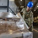 A U.S. Navy sailor in NWU Type III camouflage uniform and blue hard hat is wrapping a pallet with stretch plastic wrap inside what appears to be a large ship hangar bay or warehouse facility. The sailor is actively securing cargo on a wooden pallet using clear stretch film. A modular building unit branded 'MOBILEASE' is visible in the background along with scaffolding and overhead lighting.