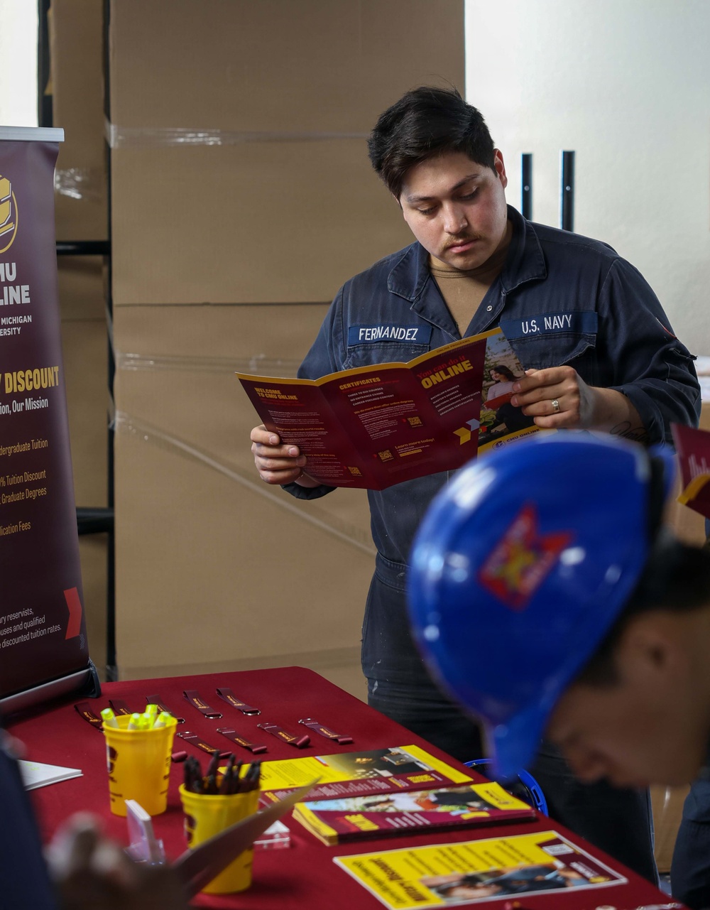 A U.S. Navy sailor named Fernandez is reading a Central Michigan University (CMU) Online brochure at what appears to be an educational fair or recruitment event. The table in the foreground is set up with CMU-branded materials in maroon and gold colors, including brochures, pens, and highlighters. A CMU Online banner is visible to the left, advertising military discounts on tuition.
