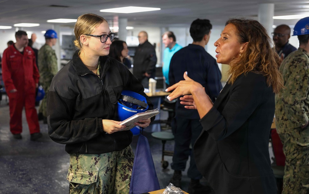 A military personnel member in U.S. Navy digital camouflage uniform and black fleece jacket is engaged in conversation with a civilian woman in a black blazer. The setting appears to be an indoor military facility or ship interior, with multiple other personnel visible in the background. The naval service member is holding a blue hard hat and documents during the discussion.