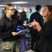 A military personnel member in U.S. Navy digital camouflage uniform and black fleece jacket is engaged in conversation with a civilian woman in a black blazer. The setting appears to be an indoor military facility or ship interior, with multiple other personnel visible in the background. The naval service member is holding a blue hard hat and documents during the discussion.