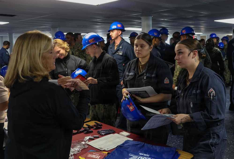 A group of U.S. Navy personnel in dark coveralls and blue hard hats are attending what appears to be an educational or career fair event, likely aboard a naval vessel or facility. Sailors are interacting with a civilian representative at a table displaying materials from what appears to be Texas Southern University (TSU). The event resembles a higher education outreach or benefits fair targeting active-duty military members.