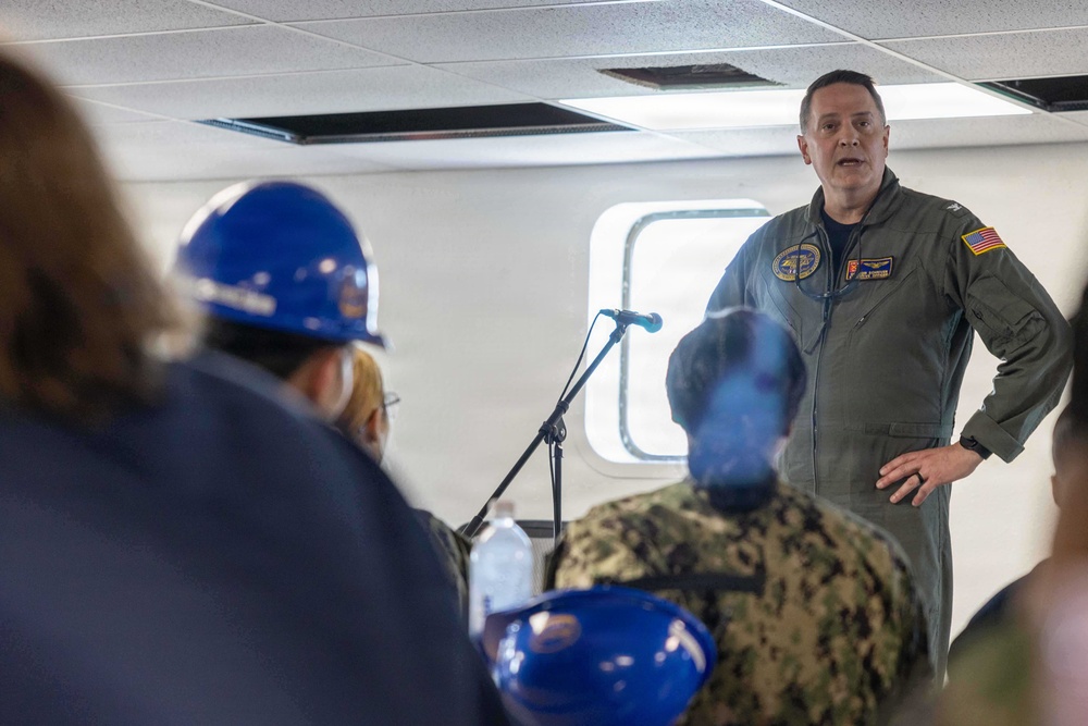 A military officer in an olive green flight suit with a U.S. flag patch is addressing a group of people in what appears to be an indoor briefing aboard a naval vessel or military facility. Several attendees are wearing blue hard hats, and at least one person in the audience is wearing a Navy digital camouflage uniform. A microphone on a stand is positioned in front of the speaker.