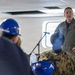 A military officer in an olive green flight suit with a U.S. flag patch is addressing a group of people in what appears to be an indoor briefing aboard a naval vessel or military facility. Several attendees are wearing blue hard hats, and at least one person in the audience is wearing a Navy digital camouflage uniform. A microphone on a stand is positioned in front of the speaker.