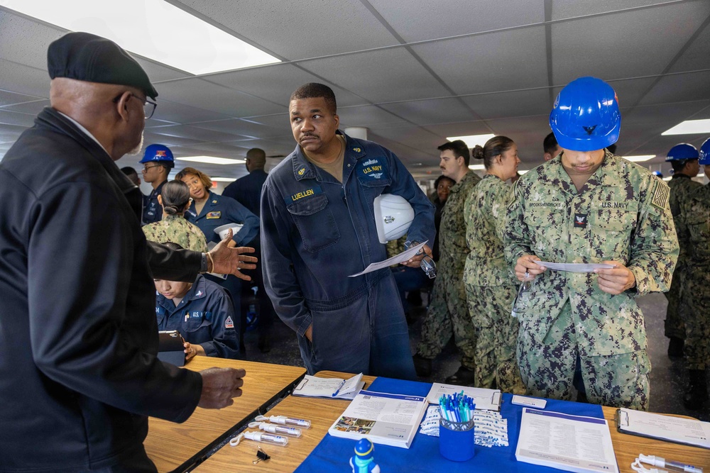 A group of U.S. Navy personnel are gathered in an indoor facility, appearing to participate in a briefing, processing event, or administrative station. A sailor in blue coveralls with the name tag 'LUELLEN' holds a white hard hat and papers while speaking with a civilian or official at a table. Other sailors in NWU Type III camouflage uniforms and blue hard hats are also present, reviewing documents.