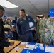 A group of U.S. Navy personnel are gathered in an indoor facility, appearing to participate in a briefing, processing event, or administrative station. A sailor in blue coveralls with the name tag 'LUELLEN' holds a white hard hat and papers while speaking with a civilian or official at a table. Other sailors in NWU Type III camouflage uniforms and blue hard hats are also present, reviewing documents.