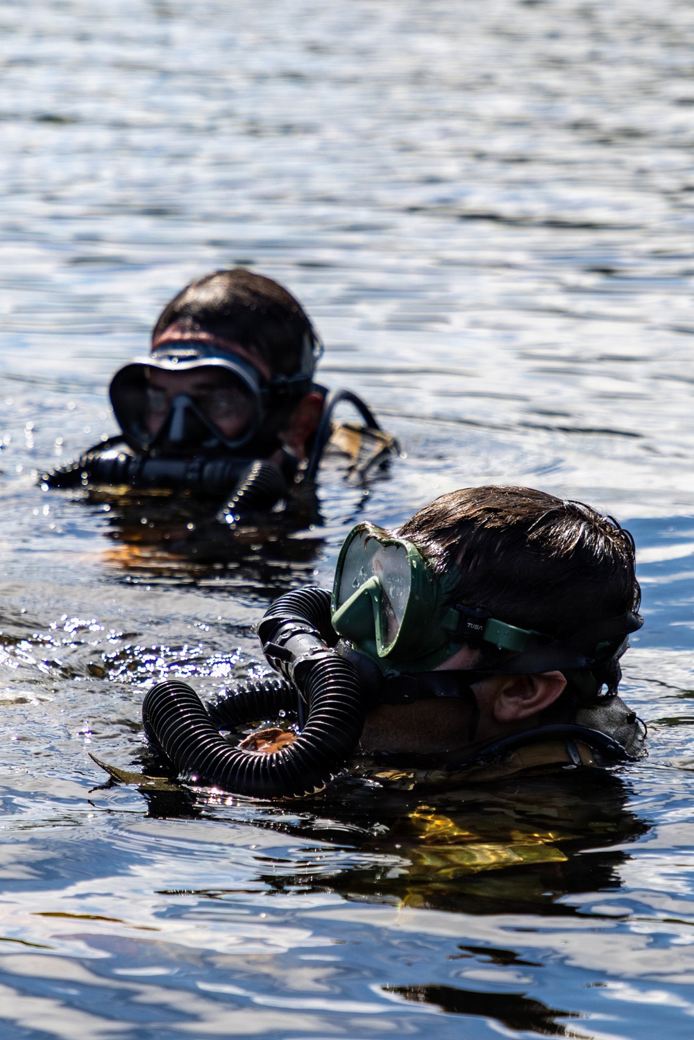 Two divers are partially submerged in open water, with only their heads and upper bodies visible above the surface. Both are wearing diving masks and wetsuits, with the foreground diver equipped with a corrugated breathing hose consistent with a rebreather or closed-circuit diving apparatus. The equipment and configuration suggest a military or tactical diving context rather than recreational diving.