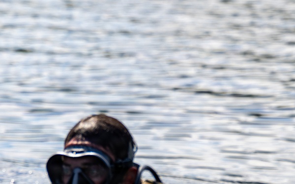 Two divers are partially submerged in open water, with only their heads and upper bodies visible above the surface. Both are wearing diving masks and wetsuits, with the foreground diver equipped with a corrugated breathing hose consistent with a rebreather or closed-circuit diving apparatus. The equipment and configuration suggest a military or tactical diving context rather than recreational diving.