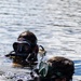 Two divers are partially submerged in open water, with only their heads and upper bodies visible above the surface. Both are wearing diving masks and wetsuits, with the foreground diver equipped with a corrugated breathing hose consistent with a rebreather or closed-circuit diving apparatus. The equipment and configuration suggest a military or tactical diving context rather than recreational diving.