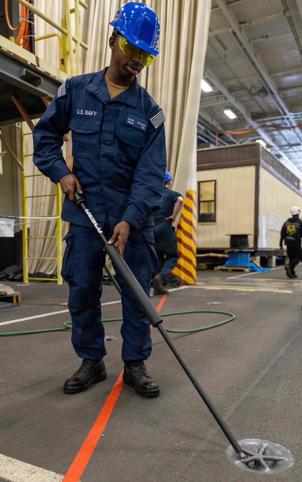 A U.S. Navy sailor in blue coveralls and a blue hard hat is operating a pressure washing or surface cleaning tool on the deck of what appears to be a naval hangar or industrial facility. The sailor is wearing safety glasses and black boots, and is using a long-handled rotary surface cleaner attached to a hose. Additional personnel and industrial equipment are visible in the background.