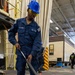 A U.S. Navy sailor in blue coveralls and a blue hard hat is operating a pressure washing or surface cleaning tool on the deck of what appears to be a naval hangar or industrial facility. The sailor is wearing safety glasses and black boots, and is using a long-handled rotary surface cleaner attached to a hose. Additional personnel and industrial equipment are visible in the background.
