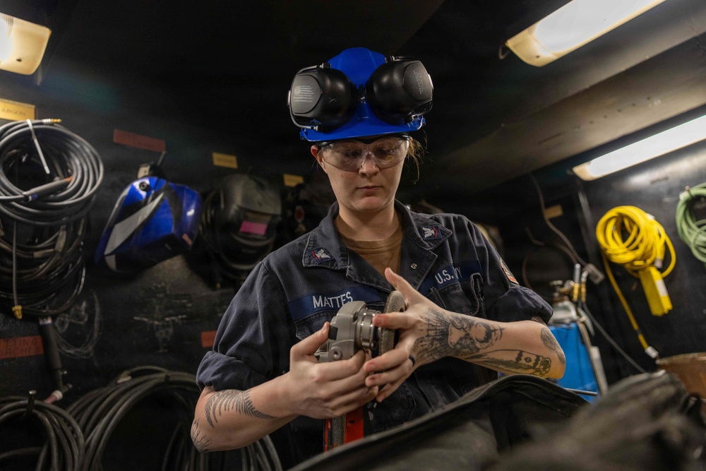 A U.S. Navy sailor named Mattes is working in what appears to be a shipboard workshop or fabrication space. She is wearing a blue hard hat with ear protection raised, safety glasses, and a navy blue coverall uniform. She is handling a metal tool or component, likely a grinder or similar equipment.