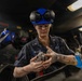 A U.S. Navy sailor named Mattes is working in what appears to be a shipboard workshop or fabrication space. She is wearing a blue hard hat with ear protection raised, safety glasses, and a navy blue coverall uniform. She is handling a metal tool or component, likely a grinder or similar equipment.
