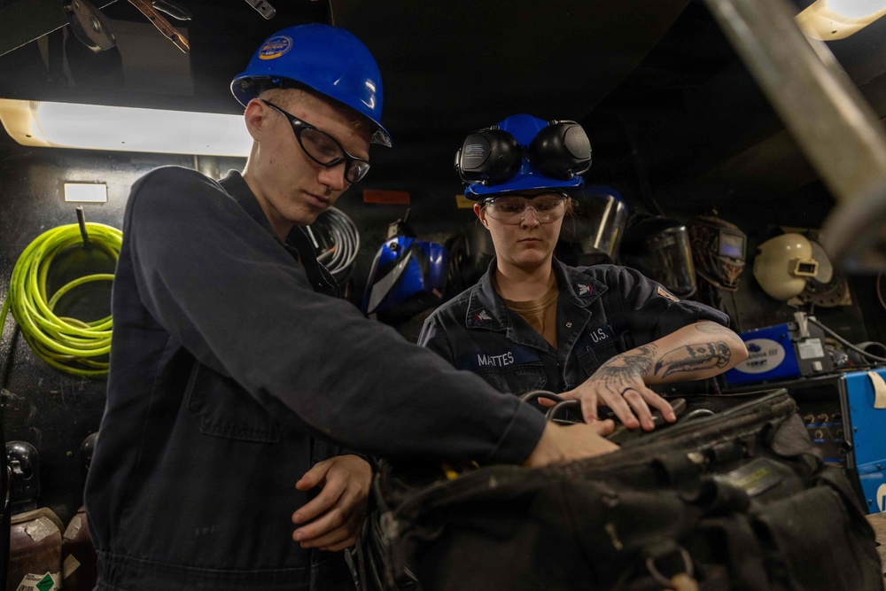 Two U.S. Navy personnel wearing blue hard hats and safety glasses are working together on mechanical equipment in what appears to be a shipboard workshop or maintenance space. The individual on the right has a name tape reading 'MATTES' and is wearing a U.S. Navy uniform with visible tattoos on their arm. Both are focused on hands-on maintenance or repair work on a piece of machinery.