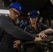 Two U.S. Navy personnel wearing blue hard hats and safety glasses are working together on mechanical equipment in what appears to be a shipboard workshop or maintenance space. The individual on the right has a name tape reading 'MATTES' and is wearing a U.S. Navy uniform with visible tattoos on their arm. Both are focused on hands-on maintenance or repair work on a piece of machinery.