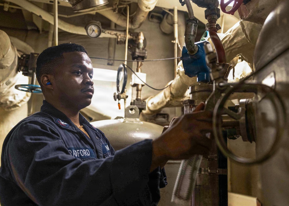 A U.S. Navy sailor identified by name tape as 'BRADFORD' is working in what appears to be a ship's engine room or mechanical space. The sailor is wearing dark blue coveralls and blue foam ear protection, and is operating or inspecting a valve or mechanical fitting. The environment is densely packed with pipes, gauges, valves, and insulated equipment typical of a naval vessel's engineering spaces.