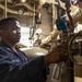 A U.S. Navy sailor identified by name tape as 'BRADFORD' is working in what appears to be a ship's engine room or mechanical space. The sailor is wearing dark blue coveralls and blue foam ear protection, and is operating or inspecting a valve or mechanical fitting. The environment is densely packed with pipes, gauges, valves, and insulated equipment typical of a naval vessel's engineering spaces.