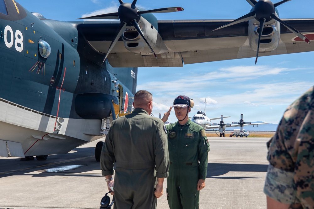 Two military aviators in green flight suits stand on an airport tarmac beside a large dark-colored military aircraft bearing the number '08' and what appears to be a Japanese roundel marking. One aviator is saluting the other in what appears to be a formal greeting or ceremony. A third person in camouflage uniform is partially visible on the right edge of the frame.
