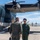 U.S. Marines, JMSDF Sailors reinforce maritime readiness during Rescue Flag 26-1 Two military aviators in green flight suits stand on an airport tarmac beside a large dark-colored military aircraft bearing the number '08' and what appears to be a Japanese roundel marking. One aviator is saluting the other in what appears to be a formal greeting or ceremony. A third person in camouflage uniform is partially visible on the right edge of the frame.