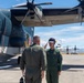 Two military aviators in green flight suits stand on an airport tarmac beside a large dark-colored military aircraft bearing the number '08' and what appears to be a Japanese roundel marking. One aviator is saluting the other in what appears to be a formal greeting or ceremony. A third person in camouflage uniform is partially visible on the right edge of the frame.