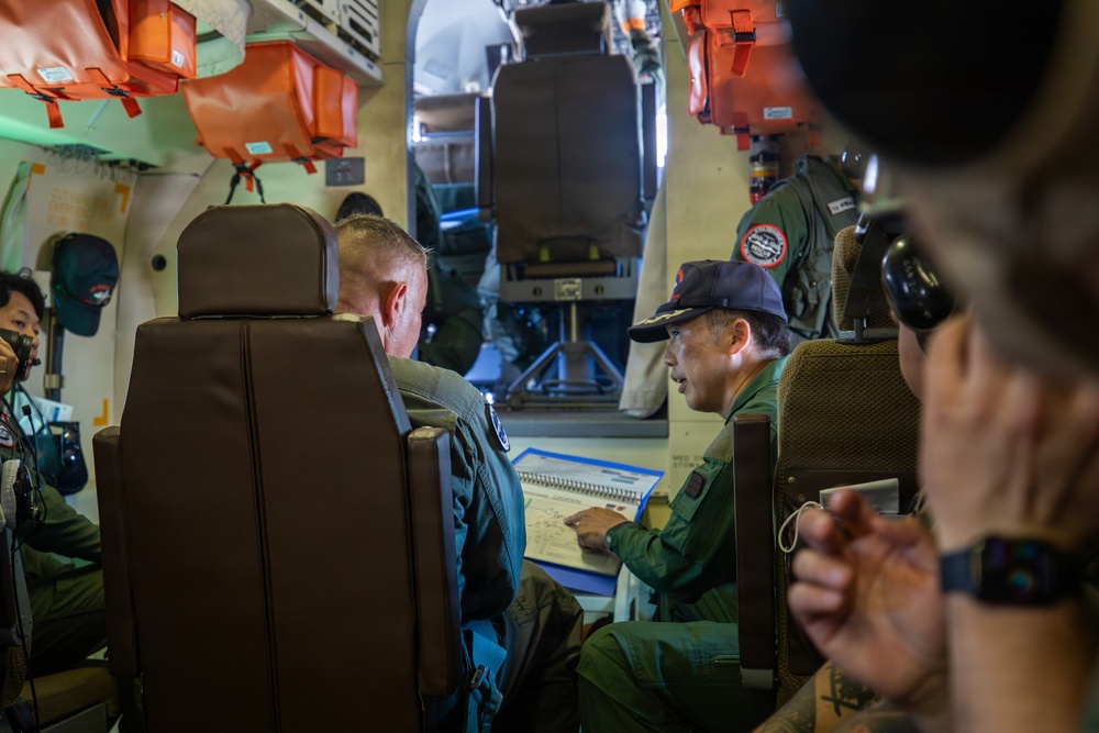 Military personnel are gathered inside what appears to be a military aircraft cabin, reviewing documents or maps together. At least two individuals in flight suits are seated and engaged in a briefing or mission planning session. Orange life vests are visible mounted overhead, consistent with a maritime patrol or transport aircraft interior.