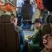 Military personnel are gathered inside what appears to be a military aircraft cabin, reviewing documents or maps together. At least two individuals in flight suits are seated and engaged in a briefing or mission planning session. Orange life vests are visible mounted overhead, consistent with a maritime patrol or transport aircraft interior.