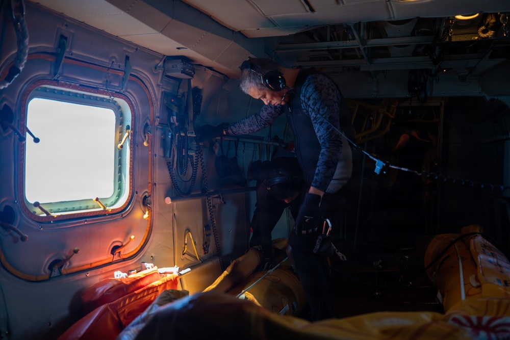 The image shows the interior of a military or search-and-rescue helicopter with at least two personnel visible. One individual wearing a camouflage uniform and communications headset is leaning over what appears to be equipment or a casualty on the floor. The scene is dimly lit with warm orange lighting accents, suggesting an active operational mission.