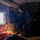 U.S. Marines, JMSDF Sailors reinforce maritime readiness during Rescue Flag 26-1 The image shows the interior of a military or search-and-rescue helicopter with at least two personnel visible. One individual wearing a camouflage uniform and communications headset is leaning over what appears to be equipment or a casualty on the floor. The scene is dimly lit with warm orange lighting accents, suggesting an active operational mission.